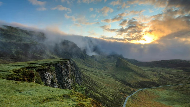 Empty paved road winding through a misty green mountain valley at sunrise, with no people, vehicles, logos, or readable text.