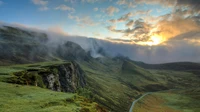 Empty paved road winding through a misty green mountain valley at sunrise, with no people, vehicles, logos, or readable text.