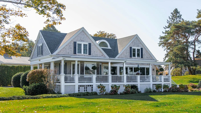 Two-story shingle house with a wraparound porch and green lawn under a clear sky, with no people, logos, or readable text.