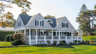 Two-story shingle house with a wraparound porch and green lawn under a clear sky, with no people, logos, or readable text.