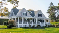 Two-story shingle house with a wraparound porch and green lawn under a clear sky, with no people, logos, or readable text.