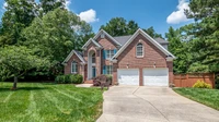 Large two-story brick house with a double garage, green lawn and mature trees under a blue sky, no people or text