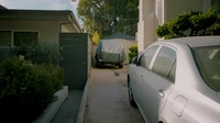 Silver sedan parked in a narrow residential driveway beside a hedge and a covered vehicle, no people or text