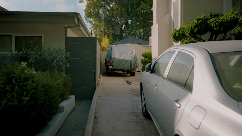 Silver sedan parked in a residential driveway beside a hedge and a covered vehicle, no people, logos, or text