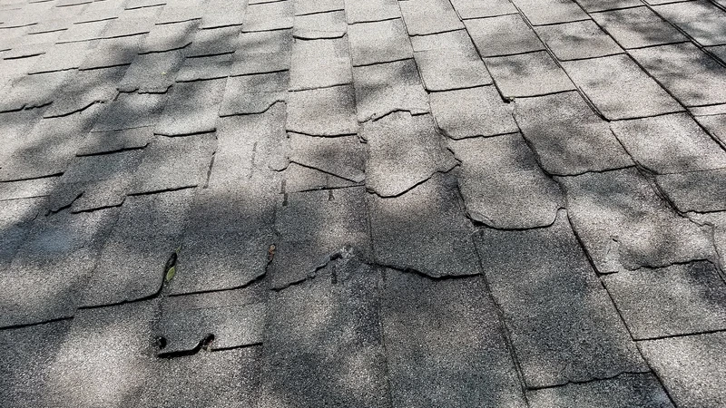 Close-up of cracked and lifted gray asphalt roof shingles after a thunderstorm