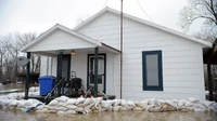 White one-story house with a row of sandbags along the front as floodwater surrounds the yard