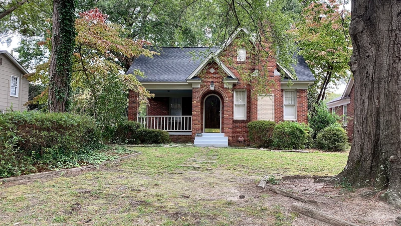 Red brick cottage-style house with a dark roof behind a tree-shaded front lawn