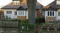 Floodwater standing in front of a row of brick and white-trimmed houses behind a wooden fence